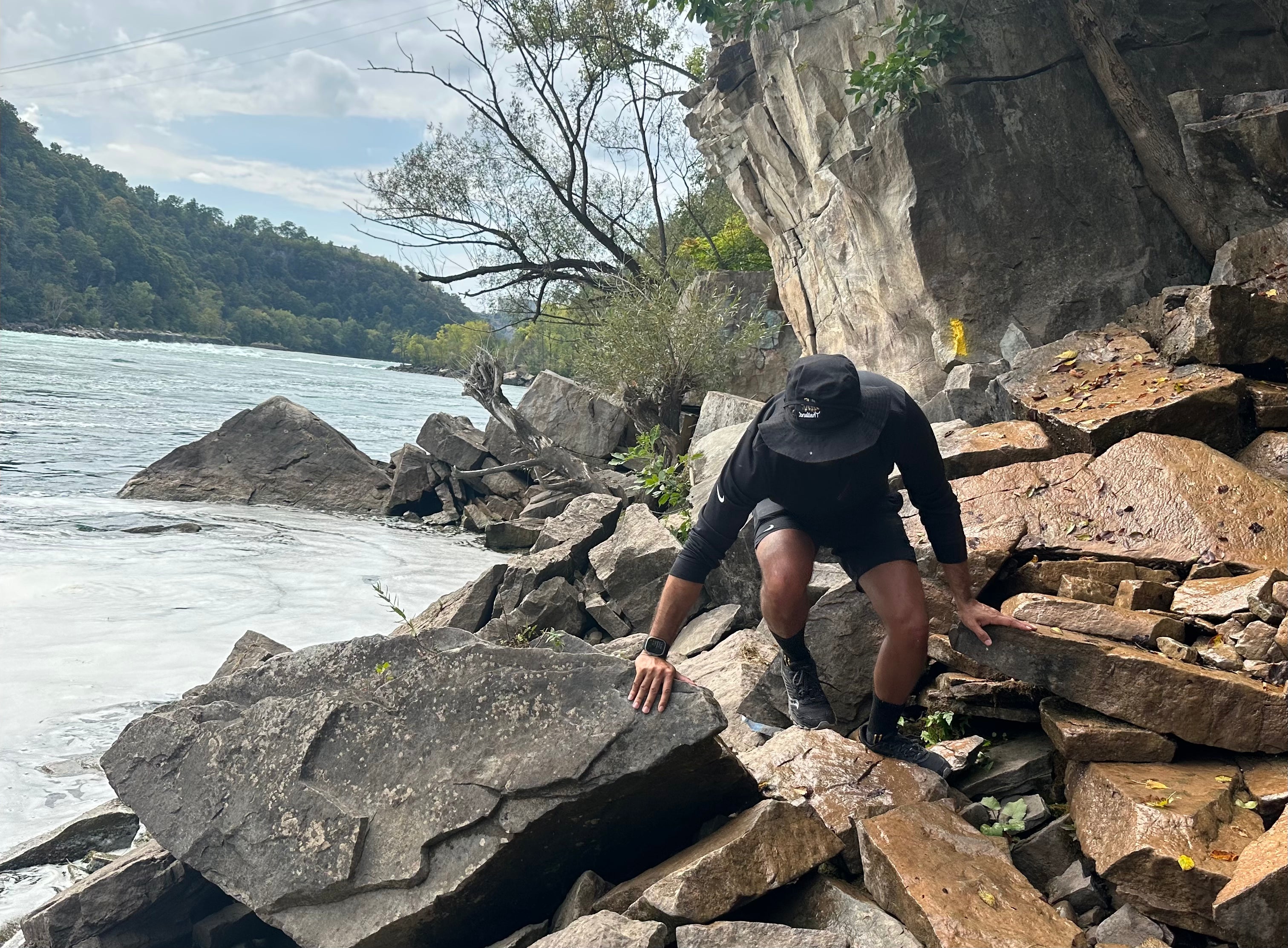 Person climbing rocky terrain by a body of water with trees and cliffs in the background